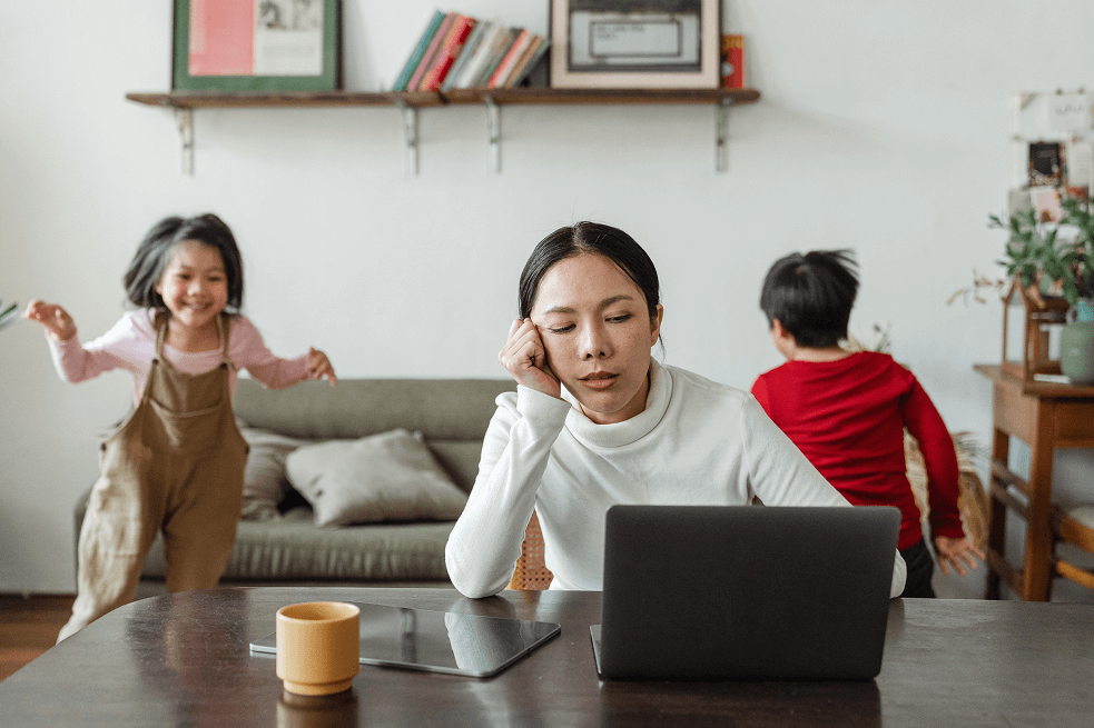 young lady with short hair sitting at her laptop with two children running behind her in the background
